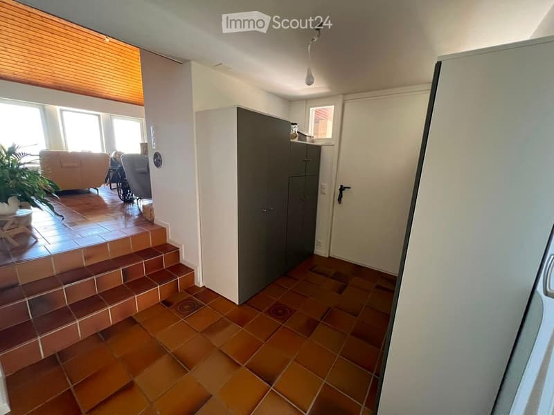 Hallway with tiled flooring, leading to a living area with a sofa, armchair, and small table. Features a staircase with tiled steps and a large window with blinds.