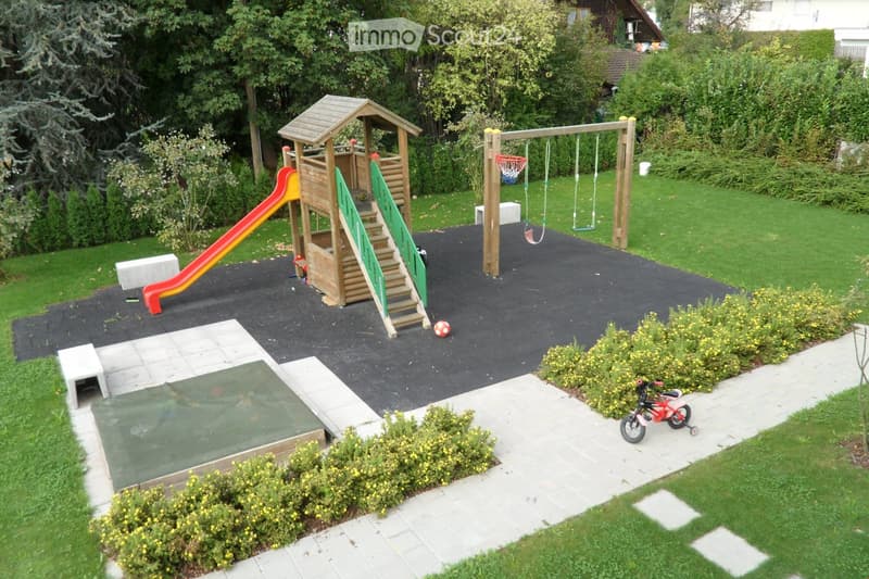 Well-maintained backyard with a wooden play structure, slide, swing set, small basketball hoop, and a covered sandbox. The area is surrounded by greenery and includes a paved pathway and small garden beds.