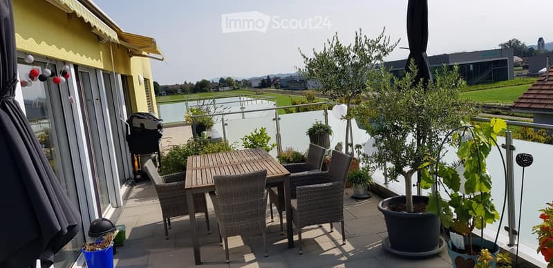 Balcony with a wooden table, wicker chairs, potted plants, and an umbrella, overlooking a scenic landscape.