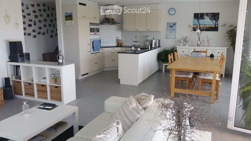 Open-plan living and dining area with a white sofa, coffee table, dining table with chairs, and a modern kitchen featuring white cabinets, an oven, microwave, and various appliances.
