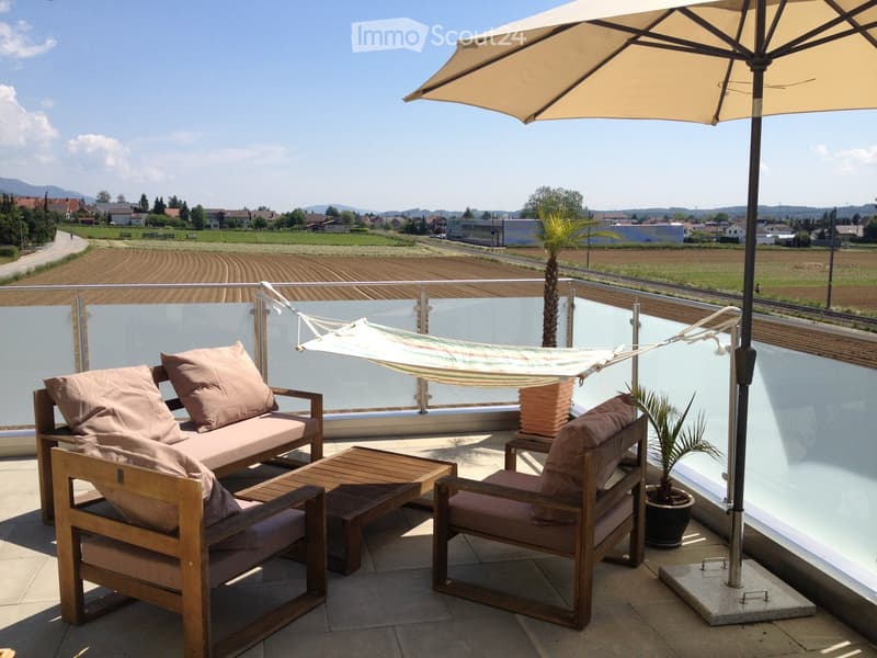 Balcony with wooden furniture, including a sofa, chairs, a small table, a hammock, and an umbrella, overlooking a scenic countryside view.