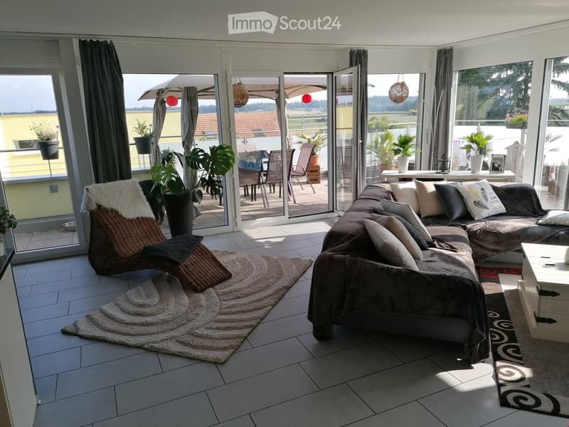 Modern living room with large glass doors leading to a balcony, featuring a sectional sofa, coffee table, wicker chair, and potted plants.