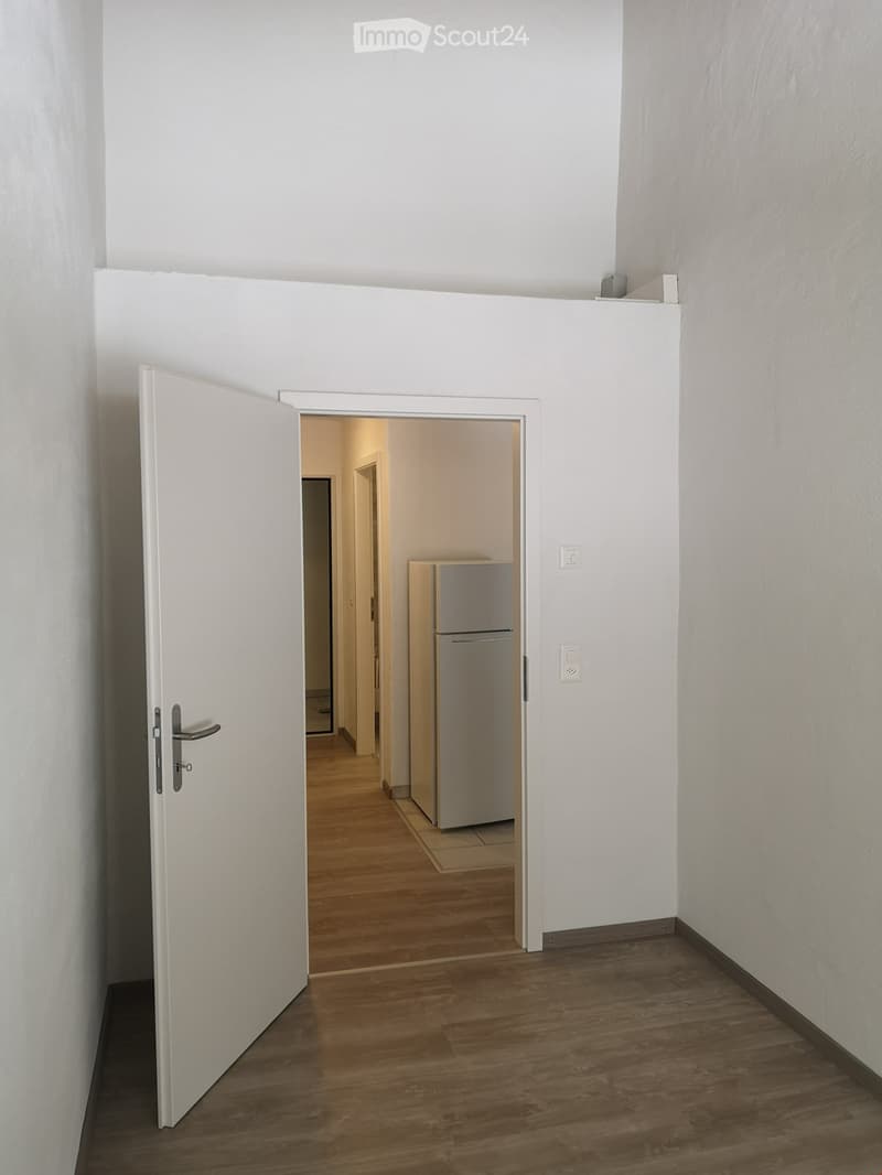 Empty hallway with wooden flooring, white walls, and an open door leading to another room with a refrigerator visible.