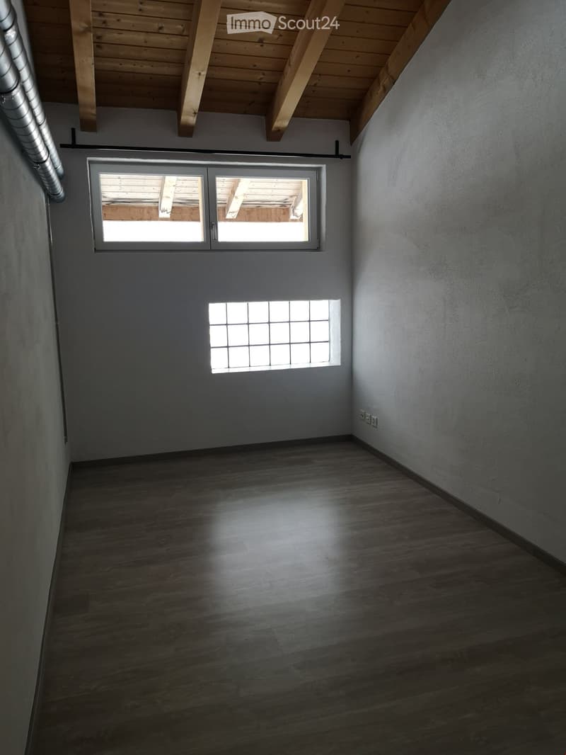 Empty room with wooden flooring, exposed wooden beams on the ceiling, and two windows allowing natural light to enter.