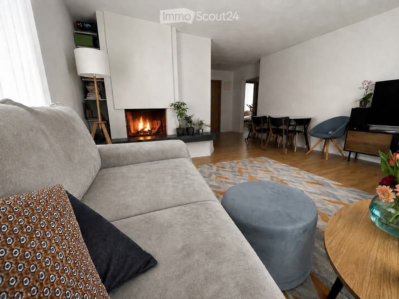 Modern living room with a grey sectional sofa, patterned throw pillow, ottoman, coffee table, floor lamp, fireplace, and dining area with chairs and table.