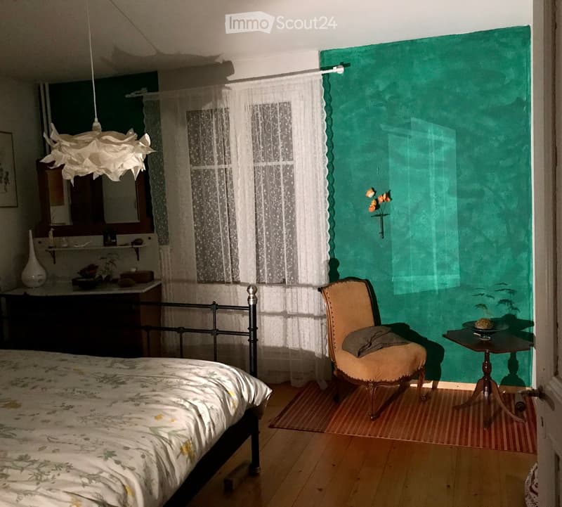 Cozy bedroom with a bed, a chair, a small table, a mirror, a sink, and a vase on a shelf. The room features wooden flooring, a green accent wall, and a hanging lamp.
