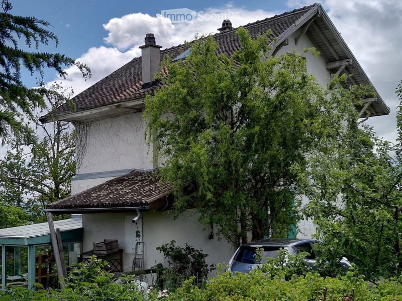 Two-story house with a tiled roof, white exterior, and overgrown garden. A car is parked in front, and there is a small wooden shed to the left.