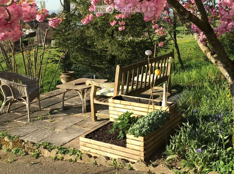 Garden area with a wooden bench, table, wicker chair, and wooden planter box with plants, surrounded by blooming trees and greenery.