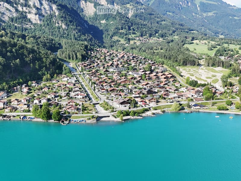 Aerial view of a picturesque village nestled in a valley surrounded by lush green forests and mountains, with a lake in the foreground.