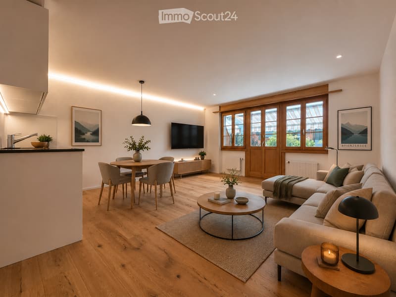 Open-plan living and dining area with light wooden flooring, a beige sectional sofa, a round coffee table, a dining table with four chairs, a hanging pendant light, and a television mounted on the wall. The space features modern decor with artwork, a side