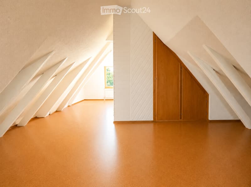 An empty attic room with exposed wooden beams, a wooden floor, and a single window with a view of greenery. The space features white walls and a large wooden door.