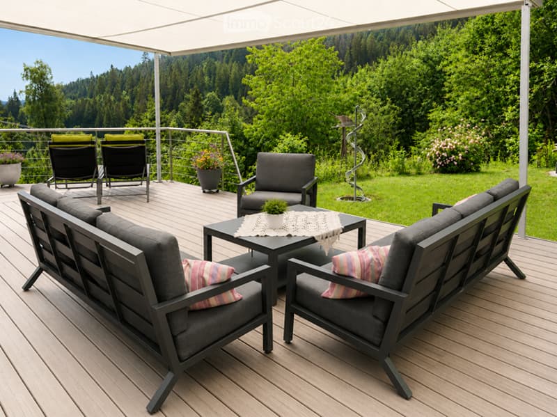 Wooden deck with grey outdoor furniture including a sofa, chairs, and a coffee table, overlooking a lush green landscape.