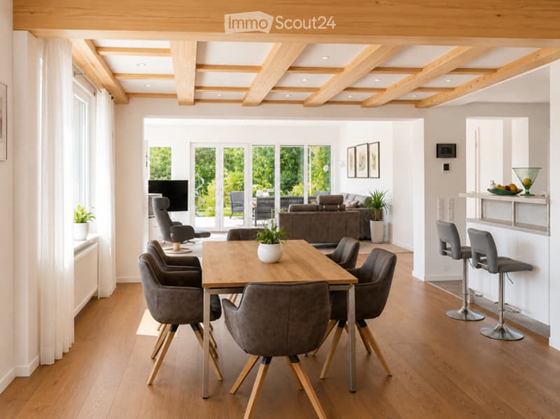 Open-plan living and dining area with wooden flooring, exposed wooden beams on the ceiling, large windows, and modern furniture including a dining table with six chairs, a sofa, and bar stools.