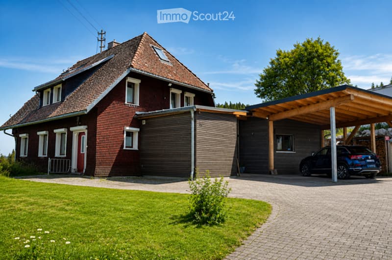 Two-story house with red brick exterior, white-framed windows, and a covered driveway leading to a garage. A modern carport shelters a parked vehicle.