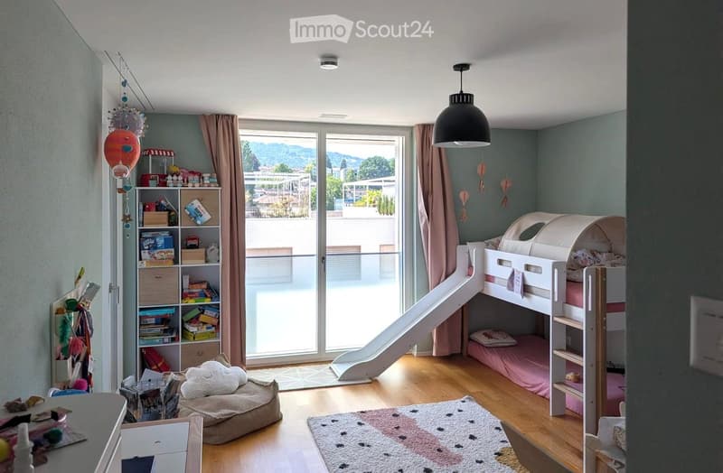 A children's bedroom with a bunk bed, slide, bookshelf, and a dog bed. The room features wooden flooring, large windows with a view, and decorative elements like hanging lanterns and a colorful rug.