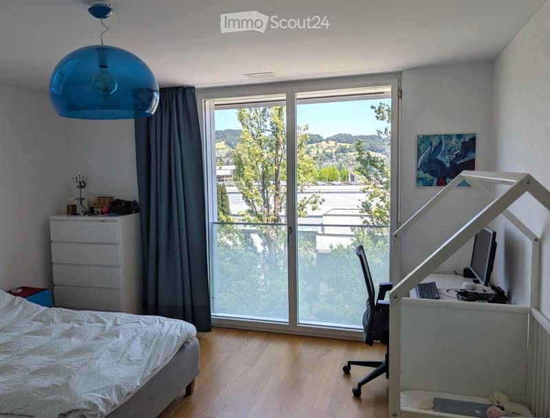 Bright bedroom with wooden flooring, large windows offering a scenic view, a bed with white bedding, a desk with a computer, and a modern hanging lamp.