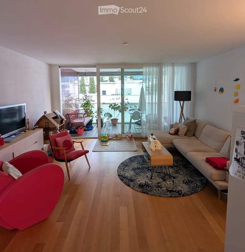 Modern living room with wooden flooring, large sliding glass doors leading to a balcony, a beige sofa, a red armchair, a coffee table, and a TV on a stand.