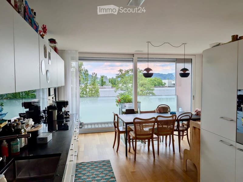 Open-plan kitchen and dining area with modern white cabinets, a black countertop, and a dining table with six chairs. The space features large windows with a view of greenery and a balcony area.