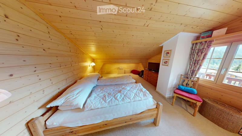 Cozy wooden bedroom with a large bed, a small chair with a cushion, a TV screen, and a window with curtains.
