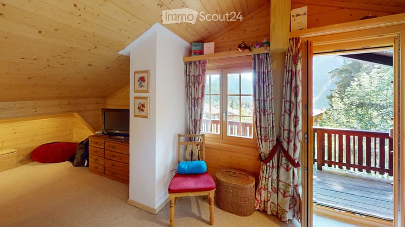 Cozy wooden bedroom with slanted ceiling, large window with view, red cushioned chair, TV, and decorative items.