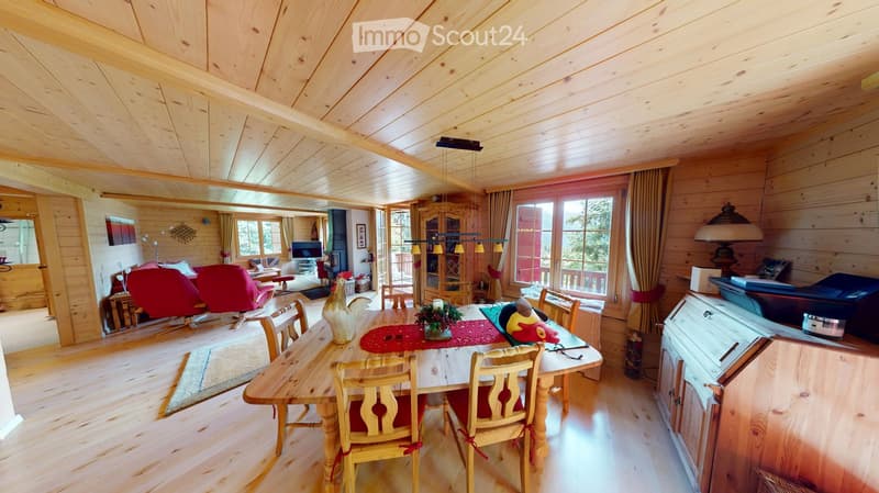 A spacious, well-lit living and dining area with wooden walls and ceiling, featuring a large dining table with chairs, a red sofa, a television, and a fireplace.