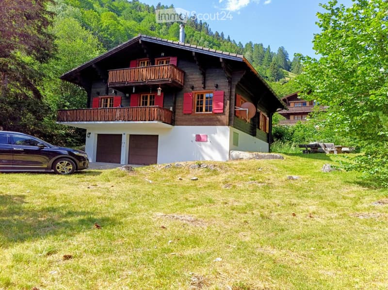 A two-story wooden house with red shutters, a balcony, and a garage, situated in a grassy area surrounded by trees.