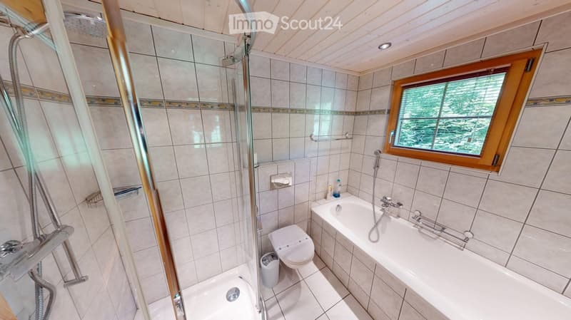 Modern bathroom with a bathtub, toilet, and walk-in shower. Features tiled walls, a window with wooden frame, and a small trash bin.