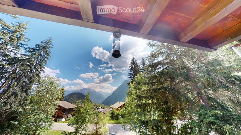 A serene outdoor view from a wooden deck, showcasing a clear blue sky, lush green trees, and distant mountain ranges.