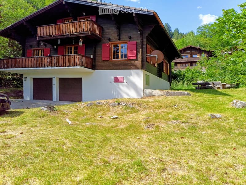 Two-story wooden house with red shutters, balconies, and a garage, surrounded by green grass and trees.