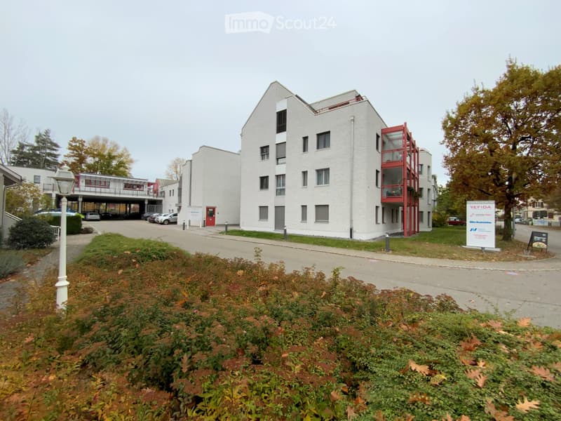 Modern multi-story building with white facade, red accents, and surrounding greenery.