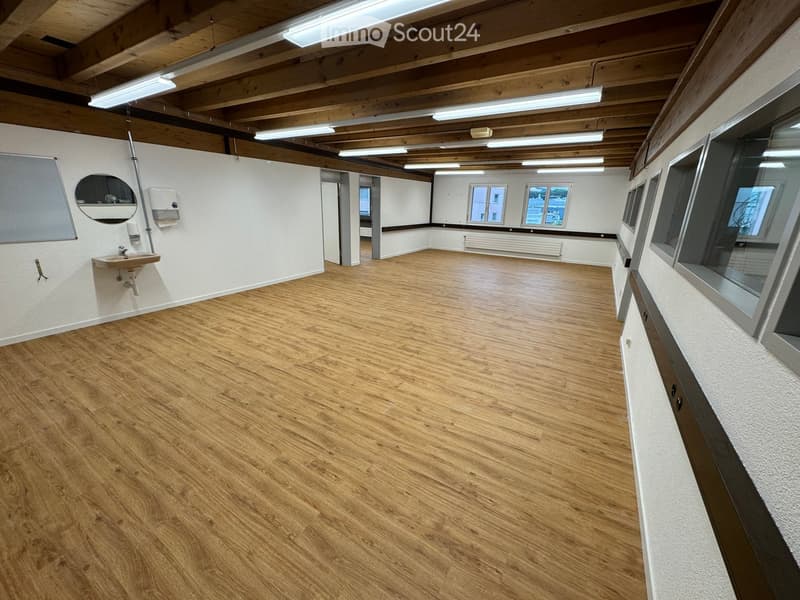 Empty room with wooden flooring, white walls, and exposed wooden beams on the ceiling. Features a small sink area with a mirror and a hand dryer, and large windows letting in natural light.