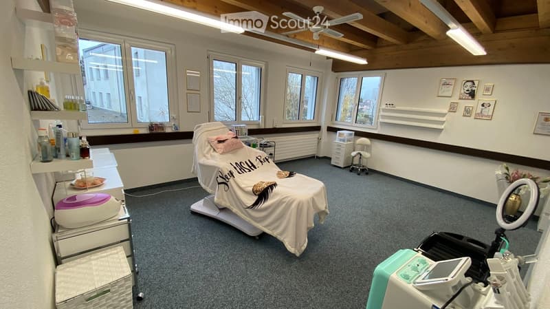 Well-lit room with wooden ceiling beams, large windows, treatment chair, shelves with cosmetic products, framed pictures, and a round mirror.