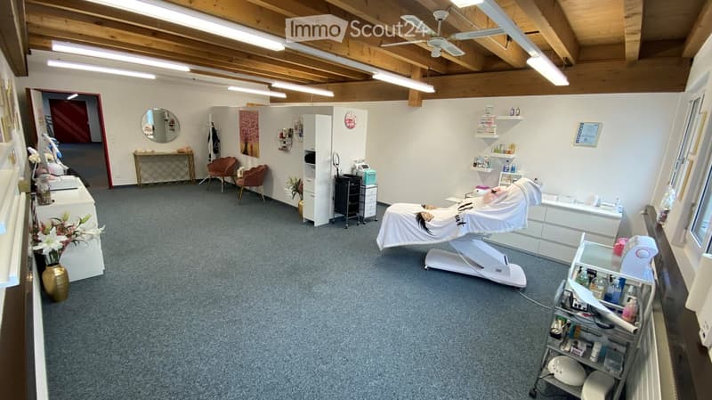 Well-lit, spacious room with exposed wooden beams and a ceiling fan. Features a treatment table, two chairs, a mirror, shelves, and decorative items.