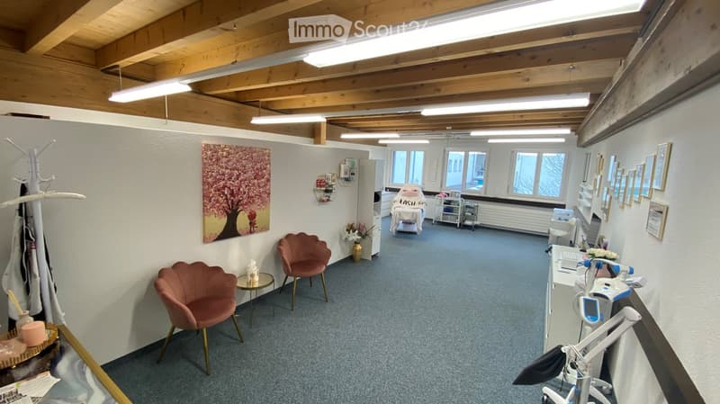 A well-lit, modern room with wooden beams on the ceiling, several chairs, a small table, a mirror, framed pictures, a vase with flowers, a cabinet, and a screen.
