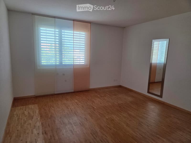 Empty room with light wooden flooring, light-colored walls, large window with blinds, sliding glass door, and a large mirror on the wall.