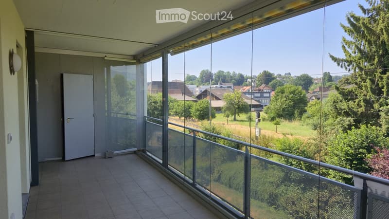 Balcony with large glass windows, tiled floor, metal railing, and a view of a green landscape with houses and trees.