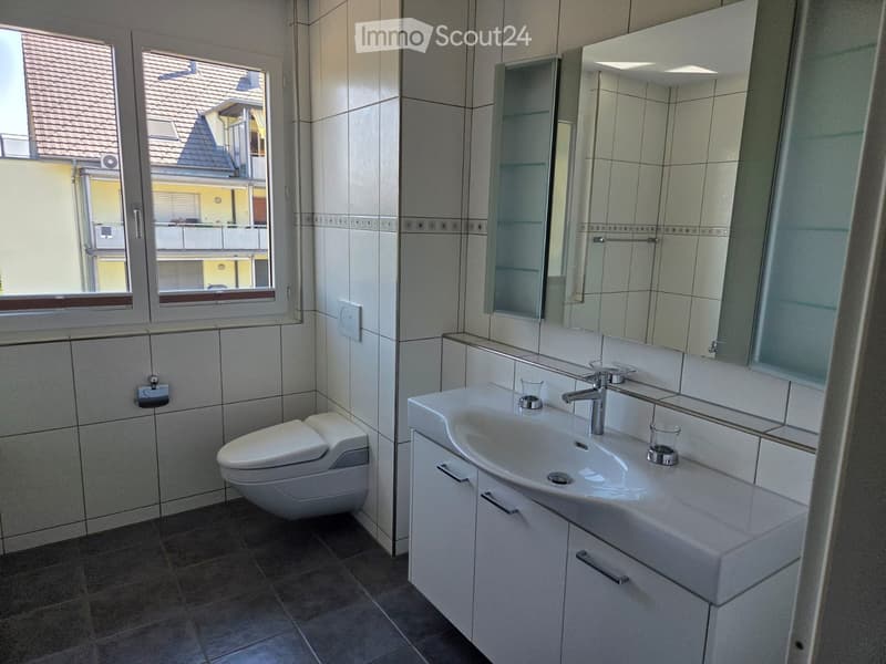 Modern bathroom with a toilet, sink, mirror, and cabinet. The room features tiled walls and a window offering a view of the building exterior.