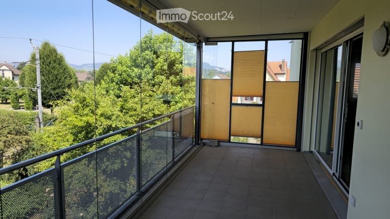 Balcony with glass railing, tiled floor, and blinds covering the windows, overlooking a green landscape with trees and houses.