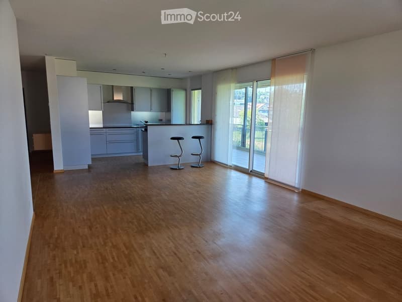 Empty room with wooden flooring, white walls, and large windows with blinds. Kitchen area visible in the background with cabinets and a countertop.