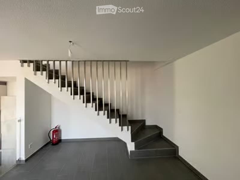 Modern hallway with a sleek, minimalist staircase featuring metal railings and dark wooden steps. The walls are white, and the floor is dark grey tiles. A red fire extinguisher is placed against the wall near the staircase.
