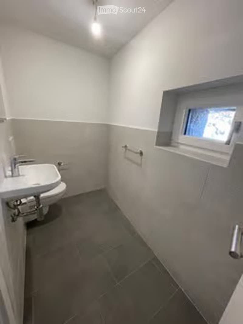 Modern bathroom with white walls, grey tiles, a window, a sink, and a toilet.