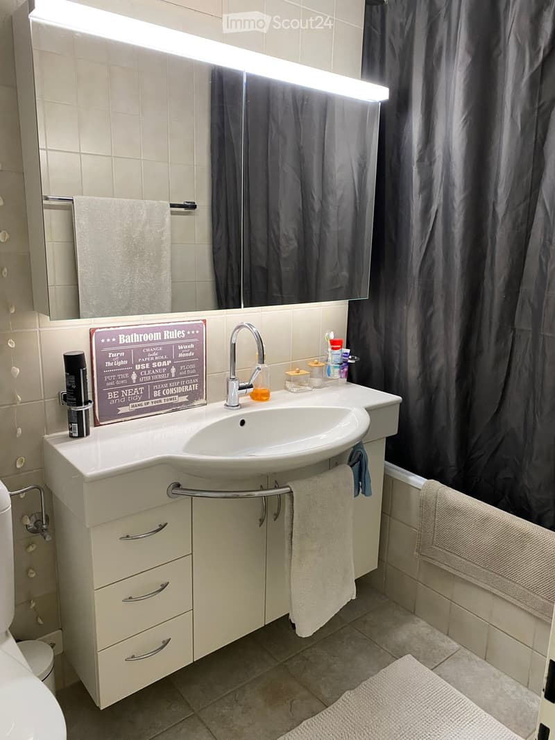 A small bathroom with a white sink, mirror, towel, cabinet, and a sign with bathroom rules. The sink has a faucet and a small shelf with toiletries. A towel hangs on a rack, and a bathroom mat is on the floor.