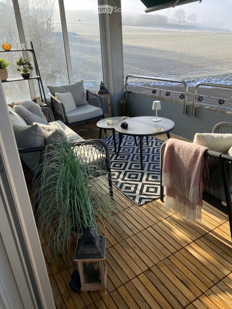 Cozy balcony with wooden decking, sofa, chairs, small tables, a lamp, a rug, and plants, overlooking a snowy landscape.