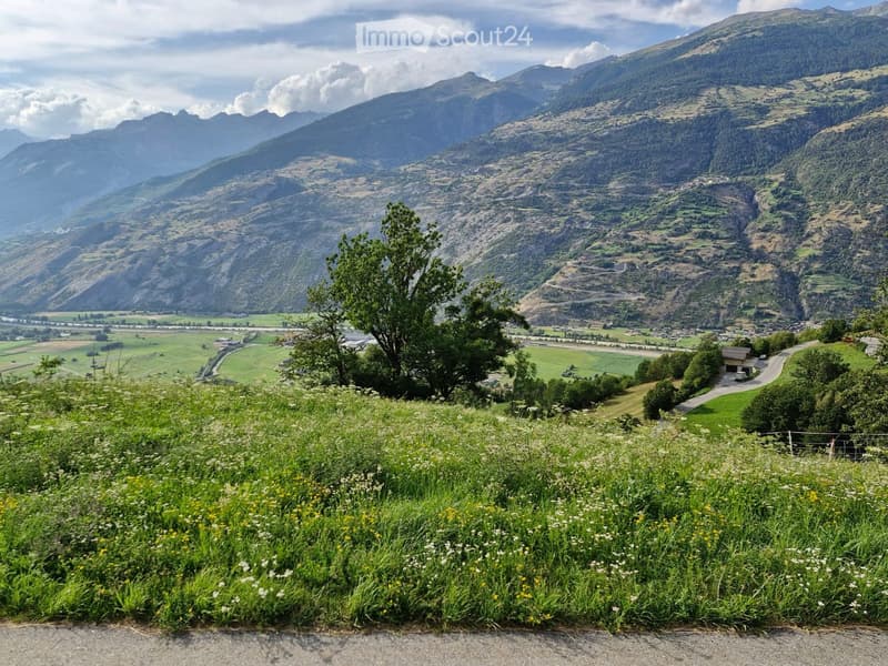 Panoramic view of a lush green valley surrounded by towering mountains, with fields, a winding road, and scattered buildings visible in the distance.