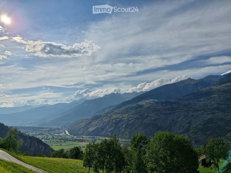 Panoramic view of a valley with mountains, green fields, and a winding river under a partly cloudy sky.