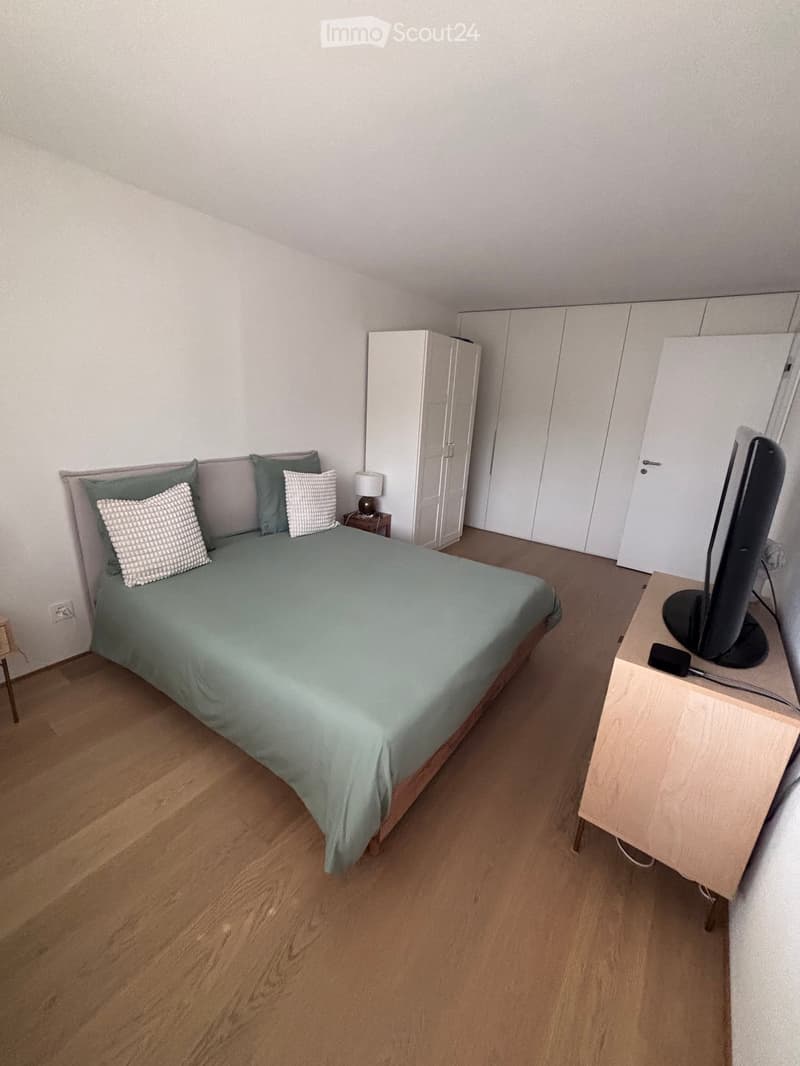 A modern bedroom with a wooden floor, a neatly made bed with green bedding and decorative pillows, a nightstand with a lamp, a wardrobe, and a desk with a computer monitor.