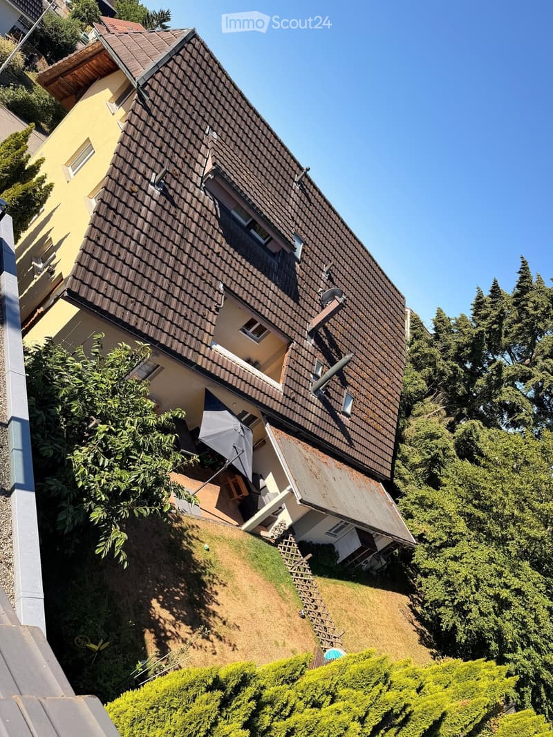 Two-story house with a steeply pitched roof and multiple windows, situated on a raised ground floor with visible steps leading up to the entrance.