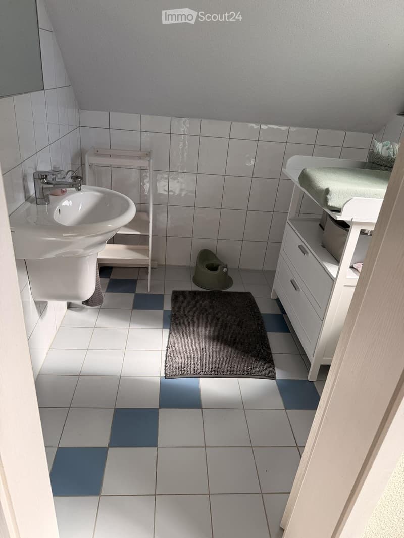 A small bathroom with white tiles, a sink with a modern faucet, a white cabinet with drawers, a mirror, a bath mat, and a small stool in the corner.