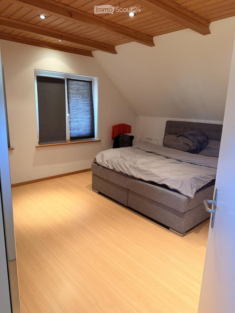 Cozy attic bedroom with wooden floors, slanted wooden ceiling, large window with blinds, and a neatly made bed.