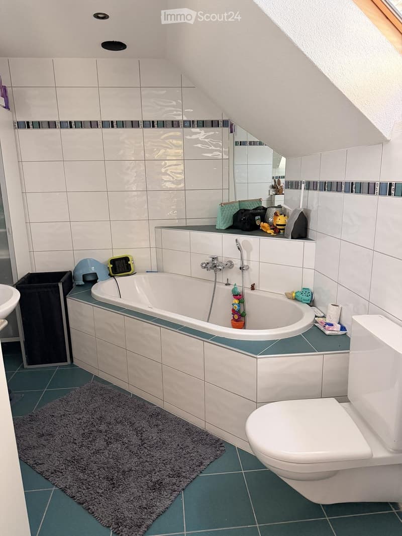 A modern bathroom with a bathtub, toilet, sink, and mirror. The walls are tiled in white with a decorative border, and the floor is green tiled. A gray bath mat is placed in front of the bathtub.